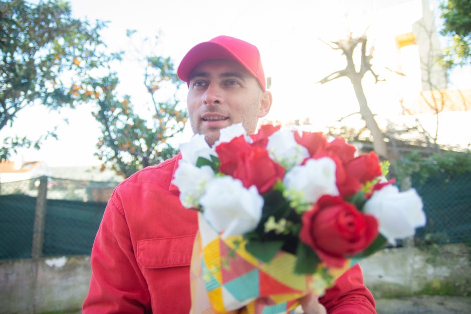 A man dressed in a red jacket and matching red cap is holding a colorful bouquet of fresh flowers, which includes red and white roses, in front of him. The bouquet is wrapped in decorative paper featu
