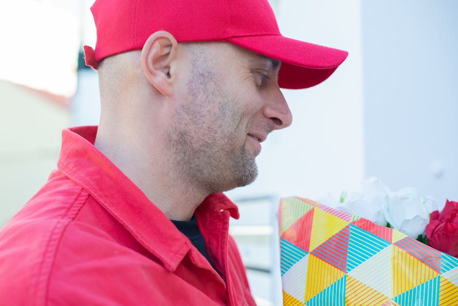 A man wearing a red cap and matching red jacket is holding a vibrant, geometrically patterned gift bag filled with white and red flowers. The arrangement showcases fresh, open petals and sturdy green 