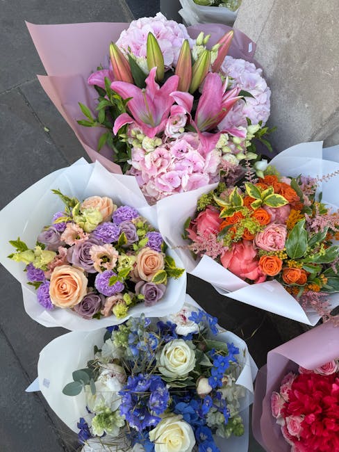 A collection of five floral bouquets displayed on a grey surface, each wrapped in elegant pastel-colored paper. The central bouquet features a prominent arrangement of pink lilies, pink hydrangeas, an