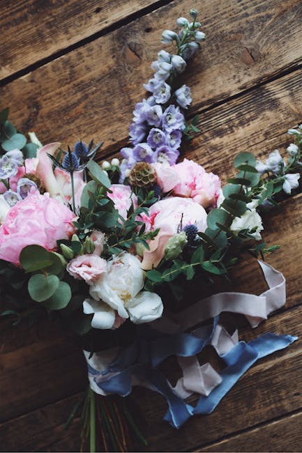 A floral bouquet featuring soft pink peonies, white and pale purple lisianthus, and sprigs of eucalyptus with rounded green leaves, arranged with dark green foliage. The bouquet is wrapped in satin ri