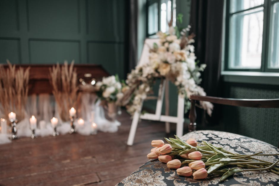 A floral arrangement featuring a variety of white and cream-colored flowers with touches of greenery, displayed on a white easel in a softly lit room with dark green walls and large windows. In the fo