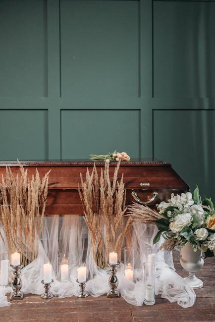 A floral arrangement placed on a wooden surface in front of a closed green paneled wall. The display includes tall, beige pampas grass and white peonies and roses arranged in a white vase, complemente