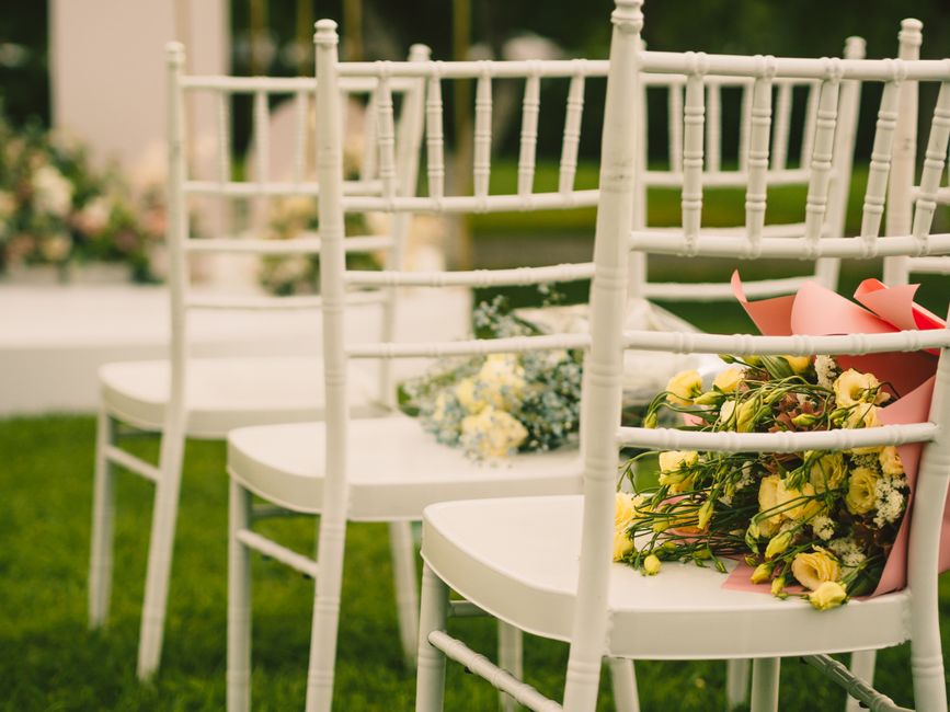 A row of white wooden chairs arranged outdoors on a grassy lawn, each with floral arrangements placed on the seats. The arrangements include yellow and white flowers such as roses and daisies, wrapped
