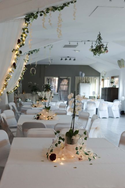 A softly lit event space decorated for a wedding at Hitchin Priory, featuring elegant floral arrangements and fairy lights. In the foreground, a white table displays a floral gift consisting of white 